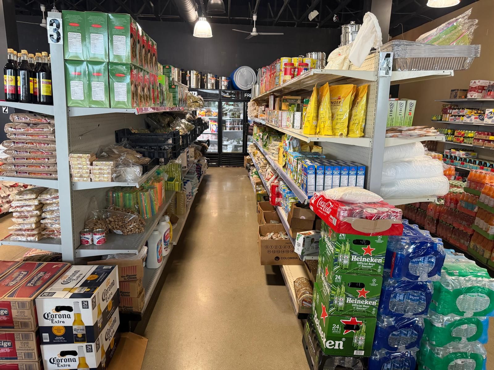 Wide view down an aisle at JNS Food Market — dried beans and grains stacked on the left, canned goods, ginger tea, and coffee on the right, with cases of Corona, Heineken, and bottled water at the end.