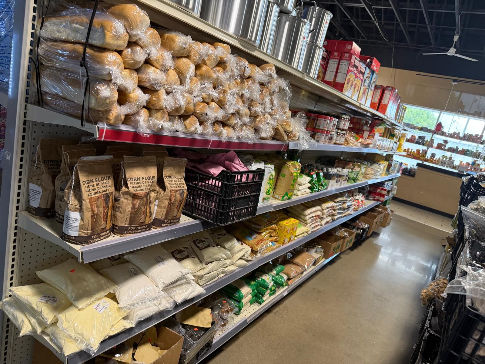 A long look down the bread and flour aisle at JNS Food Market — bagged loaves, corn flour, cassava, and bulk flour beneath a rack of aluminum stock pots.