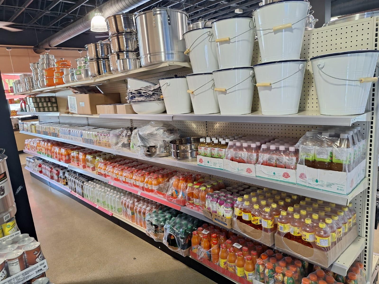 Aluminum stock pots and enamel buckets on the top rack at JNS Food Market, above shelves of tropical juice, ginger drinks, and cane syrup.