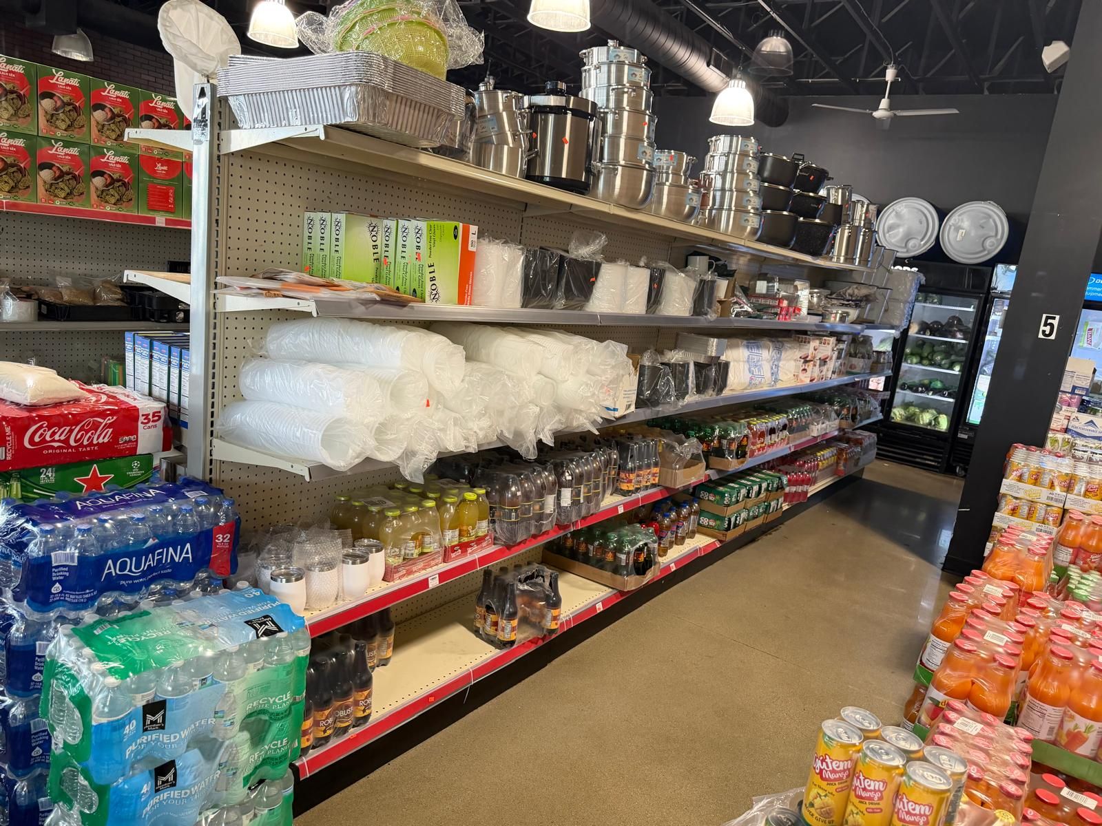 Housewares at JNS Food Market — foil pans, cookware, disposable plates and cups, and stacked cases of water and soda.