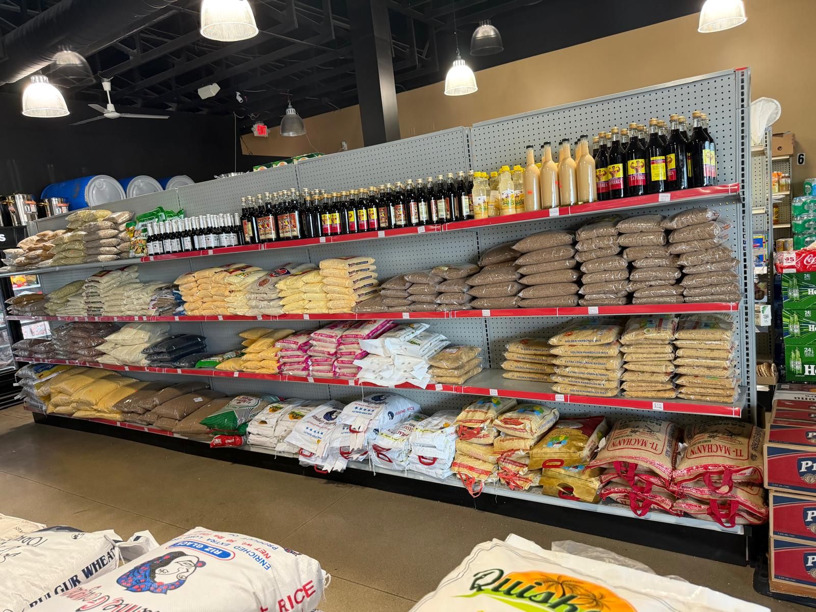 A full wall of rice and grains at JNS Food Market — sacks of jasmine rice, bulgur wheat, cornmeal, and dried beans stacked under shelves of Kola champagne and imported sodas.