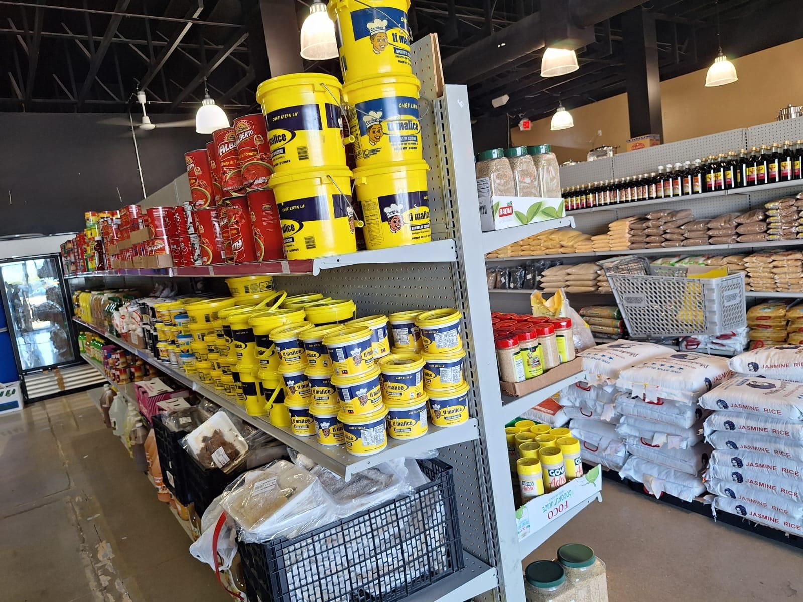 Yellow tubs of Ti Malice Haitian seasoning stacked beside canned tomatoes and tomato paste at JNS Food Market, with sacks of jasmine rice across the aisle.